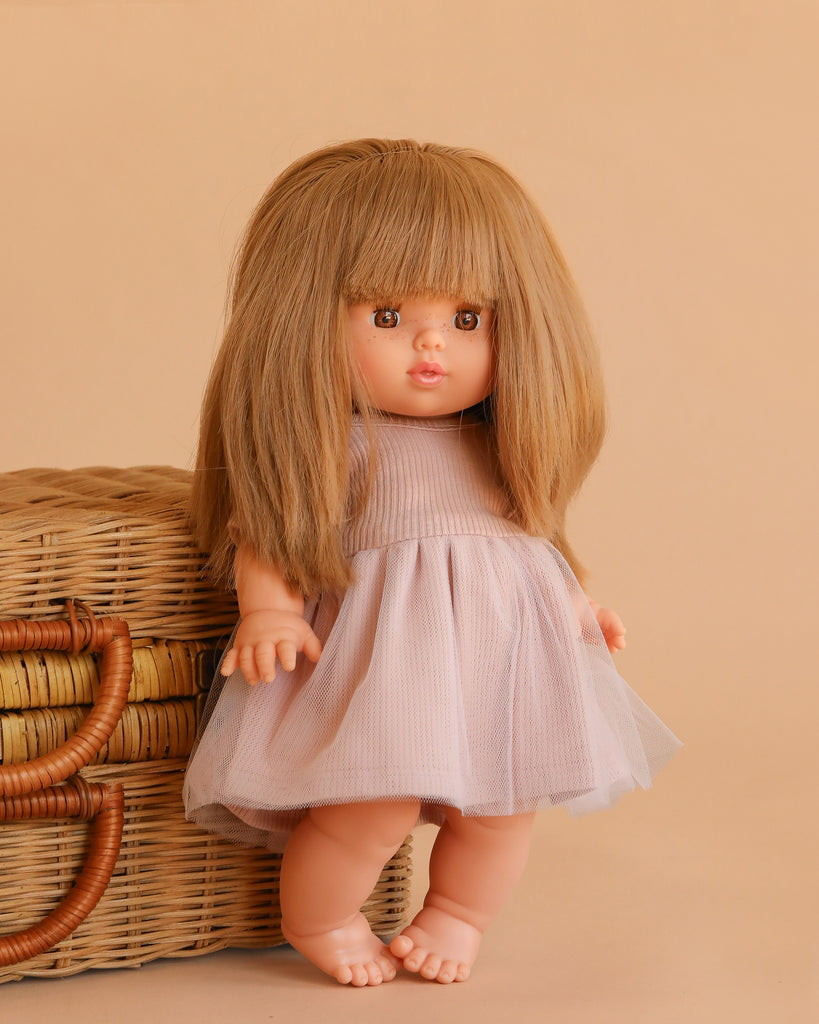 The Minikane Baby Doll (13") - Nina with Clothing has long, straight light brown hair with bangs and wears a pale pink dress. This anatomically correct doll stands barefoot by a wicker basket against a beige background.