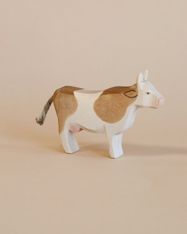 A handcrafted Ostheimer Cow - Standing with realistic brown and white patches stands against a plain beige background. The cow has painted eyes, ears, and a tail made from rope.
