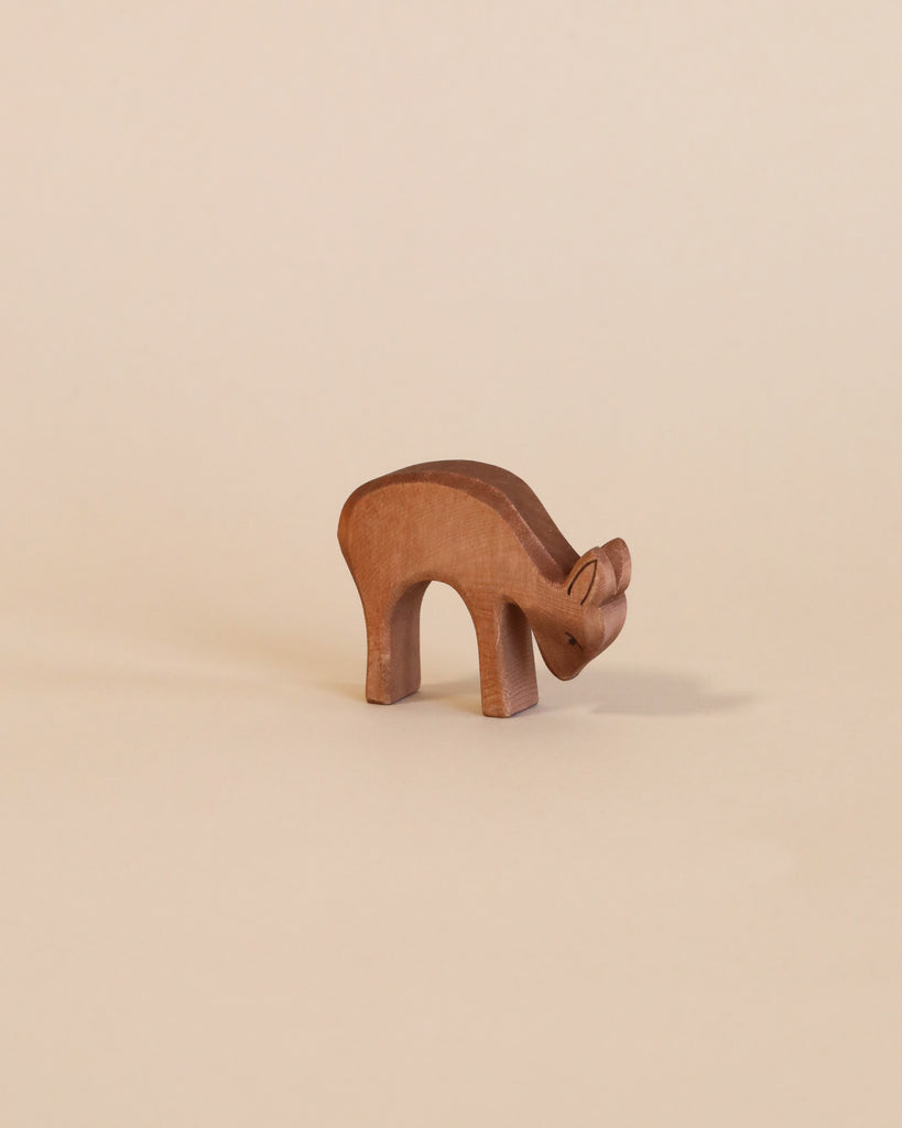 A simple Ostheimer Deer - Eating figurine on a plain beige background. This handcrafted wooden toy has a smooth, curved back and is depicted in a bent grazing pose.