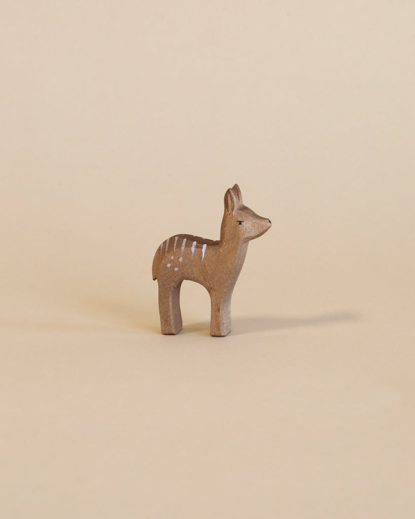 A small wooden figurine of an Ostheimer Red Deer - Fawn Standing with white spots on a solid beige background. This Ostheimer toy stands upright and is delicately carved, showcasing simple and rustic artistry ideal for imaginative play.