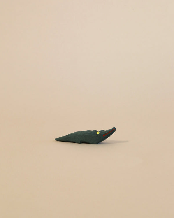 A minimalist toy crocodile, handcrafted from dark blue wood with tiny white and orange eyes, displayed against a plain light beige background.