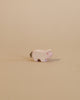A small, light-colored animal figurine from the Ostheimer Handmade Wooden Farm Animals Set stands on a beige surface with a matching background; this handcrafted toy features a short tail and subtle red accents on its face.