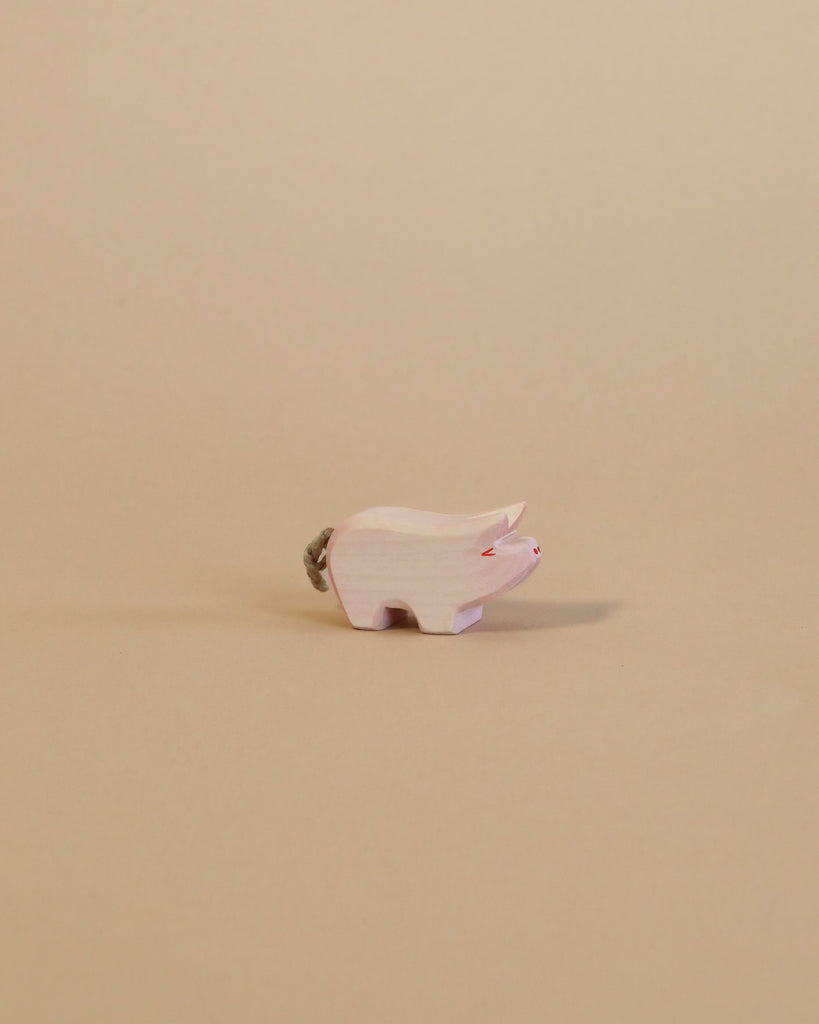 A small, light-colored animal figurine from the Ostheimer Handmade Wooden Farm Animals Set stands on a beige surface with a matching background; this handcrafted toy features a short tail and subtle red accents on its face.