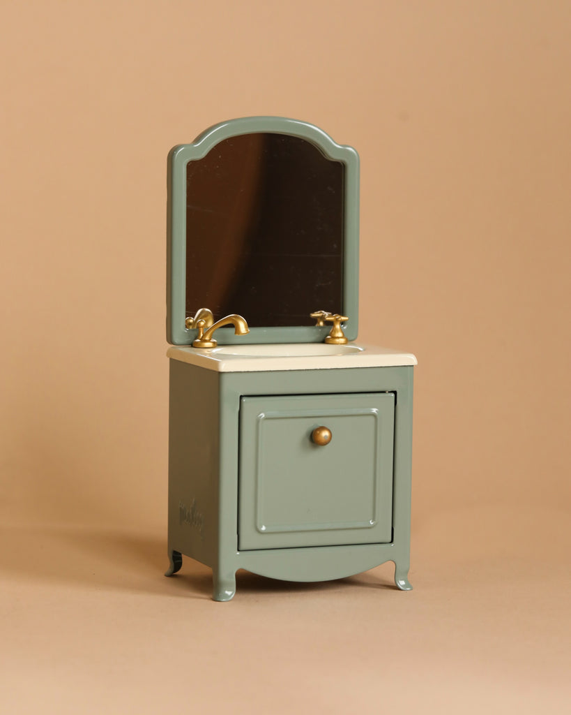 The Maileg Bathroom Starter Set features a compact, mint-green vanity with a white countertop, accented by a rectangular mirror framed in matching mint-green. The vanity includes a single cabinet door with a brass knob and is equipped with vintage-style brass faucet and handles—perfect for any Maileg little sister's cozy space.