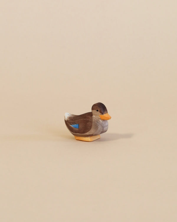 A small handcrafted Ostheimer Duck - Sitting with a predominantly gray body, an orange beak, and a distinctive blue patch on its wing, stands on a plain beige background.