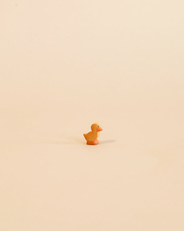 A small, orange Ostheimer Duckling centered on a plain, light beige background. The duck casts a soft shadow behind it, emphasizing its isolated position and evoking the charm of handcrafted wooden toys.