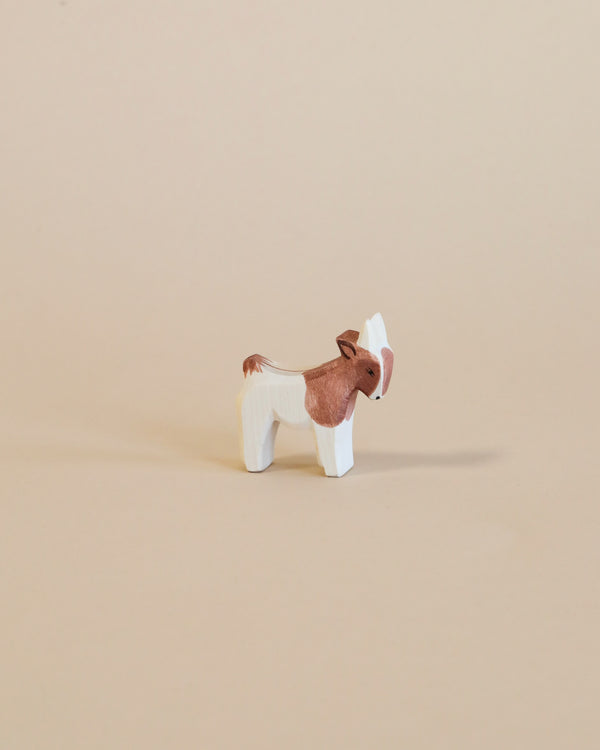 A small handcrafted Ostheimer Goat Kid - Standing with a brown body and white face and legs, standing against a plain beige background.