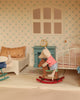 A toy mouse in a red-striped shirt and blue shorts sits on a rocking horse in the Maileg Fully Furnished Dollhouse Set + Gift With Purchase, featuring a bed, teal clock on a table, and patterned wallpaper.