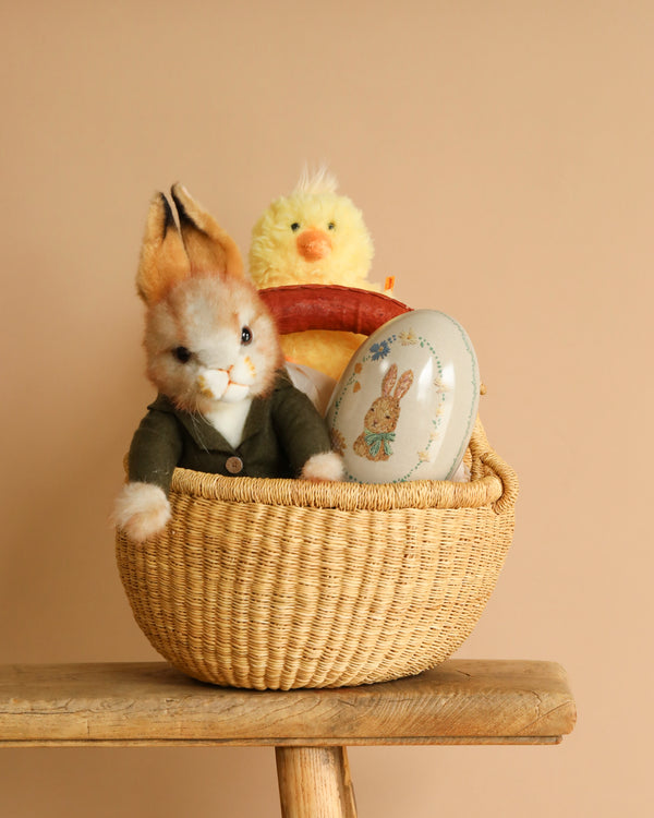 An Easter Basket Set graces a wooden bench, containing a plush toy rabbit in a green jacket, a plush yellow chick, and an egg with a rabbit illustration. The warm beige backdrop highlights the handmade charm of the scene.