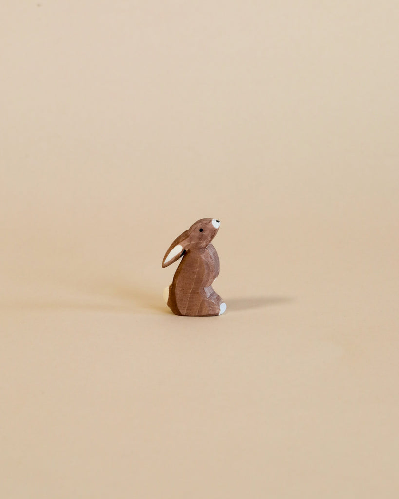 A small handcrafted Ostheimer Rabbit - Ears Low figurine stands alone against a solid, light tan background, its dark eyes and carved details facing slightly to the left.