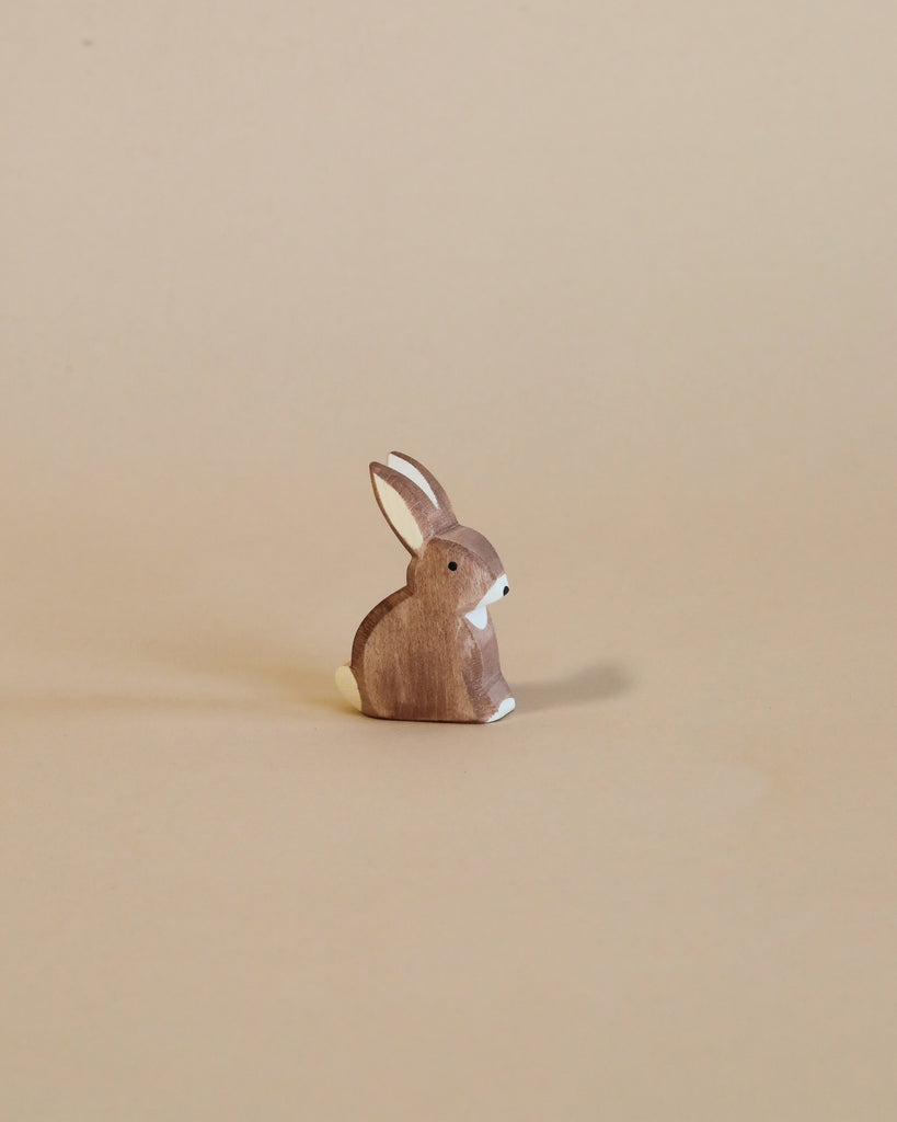 A small, brown handcrafted bunny from the Ostheimer Handmade Wooden Woodland Animals Set with white details sits on a plain beige background.