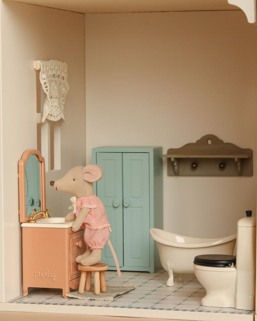 A toy mouse in pink pajamas stands at a vanity in the Maileg Fully Furnished Dollhouse Set + Gift With Purchase, featuring a bathtub, toilet, blue cabinet, shelf, and tiled floor in the dollhouse bathroom.