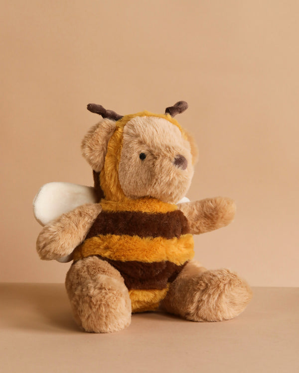 The MON AMI Bee Bear Plush Toy features a plush bear in a fuzzy yellow and brown bee costume with white wings and brown antennae, sitting on a beige surface against a matching beige background.