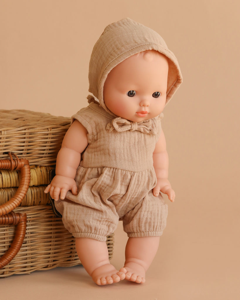 The Minikane Vintage Baby Boy Doll (13") - Louis with Clothing stands beside a wicker basket, both set against a beige background.