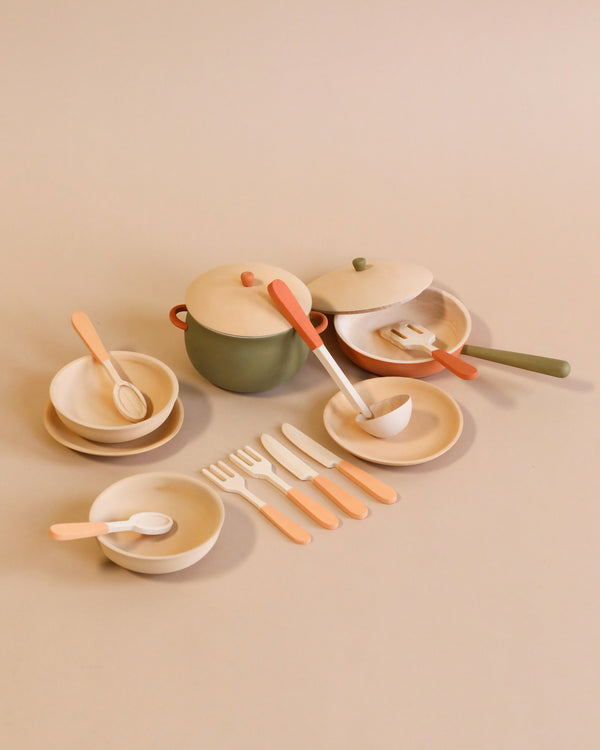 A neatly arranged set of pastel-colored non-toxic Handmade Wooden Kitchen Essentials - Herbal, including plates, a pot with a lid, utensils, and cups, set against a soft beige background.