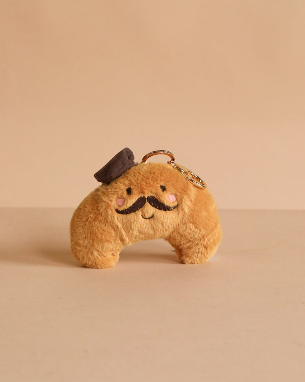 The Monsieur Croissant Keychain / Bag Charm is a plush accessory shaped like a round character with a mustache, pink cheeks, and a small brown hat, shown on a beige background.
