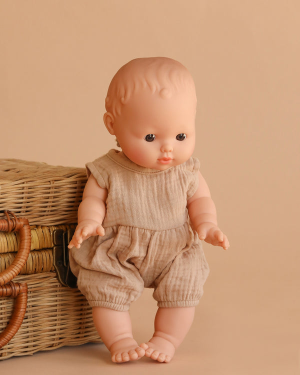The Minikane Vintage Baby Boy Doll (13") - Louis with Clothing, featuring light skin and short molded hair in a beige romper, is displayed beside a woven wicker basket on a beige background.