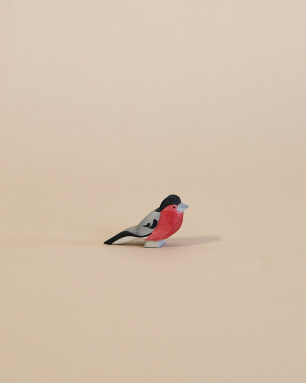 A small, colorful handcrafted Ostheimer Bullfinch with a pinkish-red chest and grey-black wings, standing isolated against a plain, light beige background.