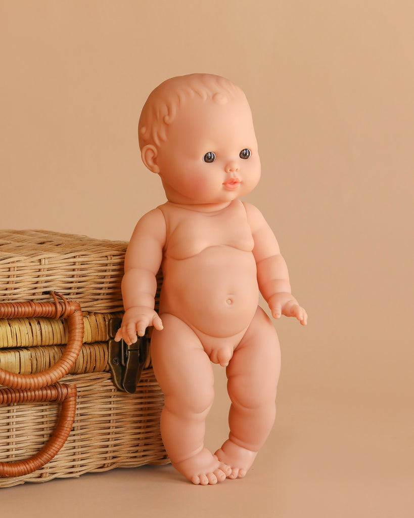 The Minikane Vintage Baby Boy Doll (13") - Louis with Clothing, featuring blue eyes and short molded hair, is displayed next to a woven wicker basket against a beige background.