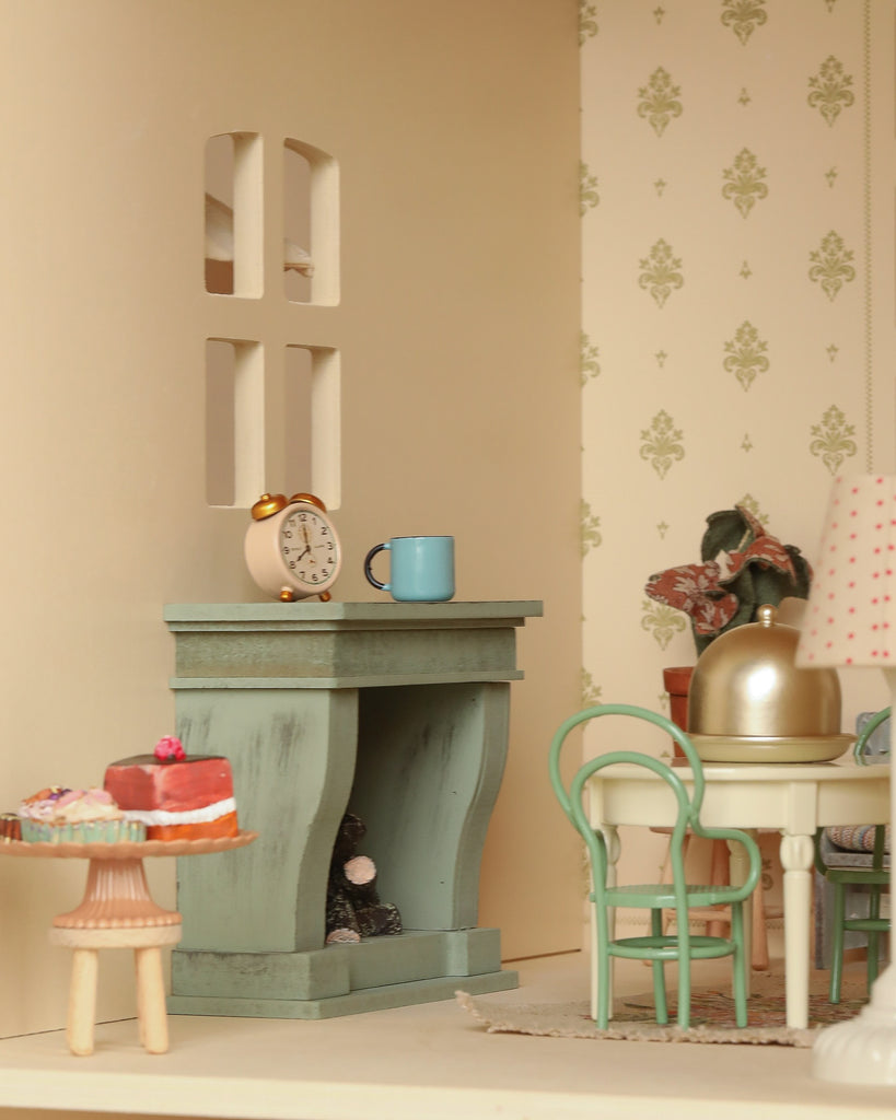 The Maileg Fully Furnished Dollhouse Set + Gift With Purchase features a cozy dining room with a green fireplace, blue mug, mantle clock, round table with chairs, cake on a stand, and floral wallpaper for a charming miniature scene.