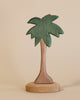 A handcrafted Ostheimer Palm Tree with Stand featuring a green crown and brown trunk, standing on a circular wooden base against a plain beige background.