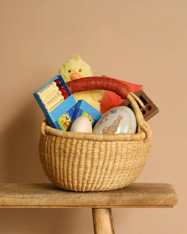 The Easter Basket Set, crafted by African villagers, includes a woven basket on a wooden bench that holds a stuffed yellow chick, chocolate bars, colored pencils, decorated eggs, and a small book. Set against a neutral beige background, it beautifully combines artistry and tradition.