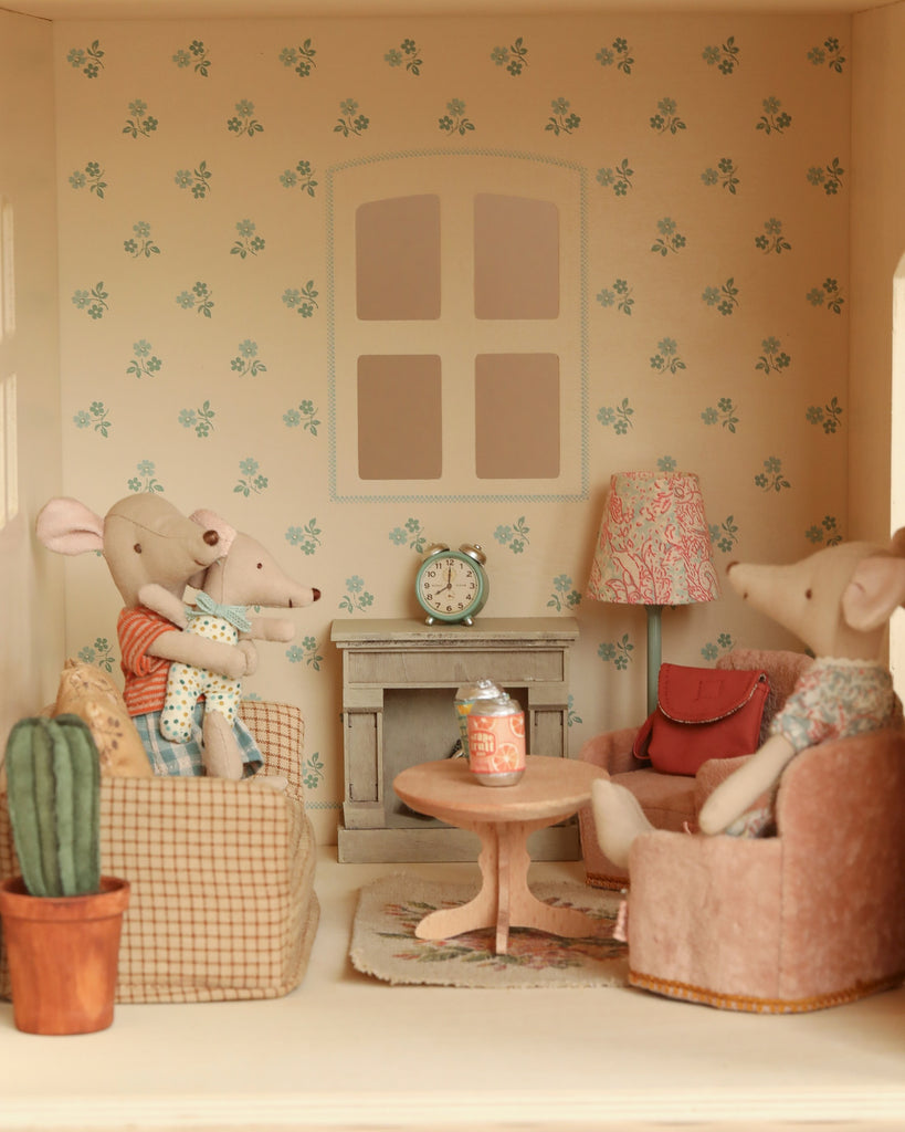 Three toy mice relax in a Maileg Fully Furnished Dollhouse Set + Gift With Purchase, complete with floral wallpaper, fireplace, clock, potted cactus, round table, and can. Two sit on the couch (one holding a baby) as another lounges in an armchair.