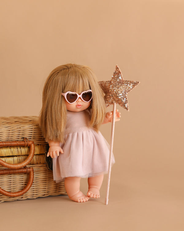 The Minikane Baby Doll (13") - Nina with Clothing and Accessories stands next to a wicker basket, wearing heart-shaped sunglasses and a pink dress while holding a sparkly star wand against a beige background.
