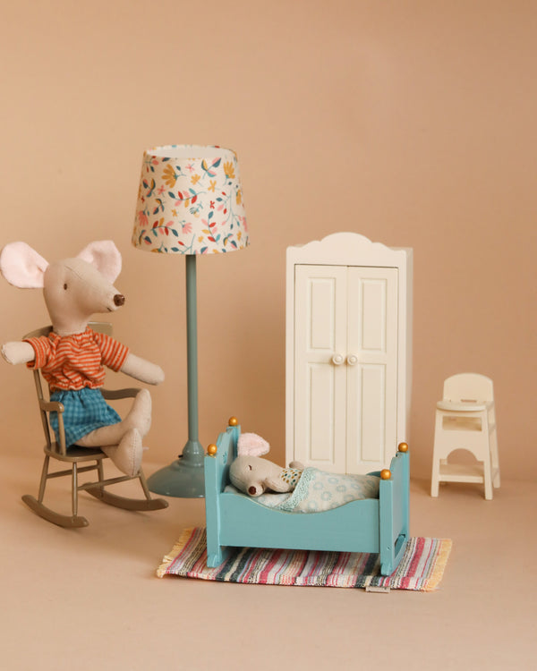 A cozy scene featuring Maileg fabric mice toys: one Mom Mouse in a rocking chair wearing a striped shirt and shorts, and one baby mouse snuggled into a blue bed. The setting includes elements from the Maileg Nursery Room Starter Set, such as a floral lamp, white wardrobe, small chair, and colorful striped rug.