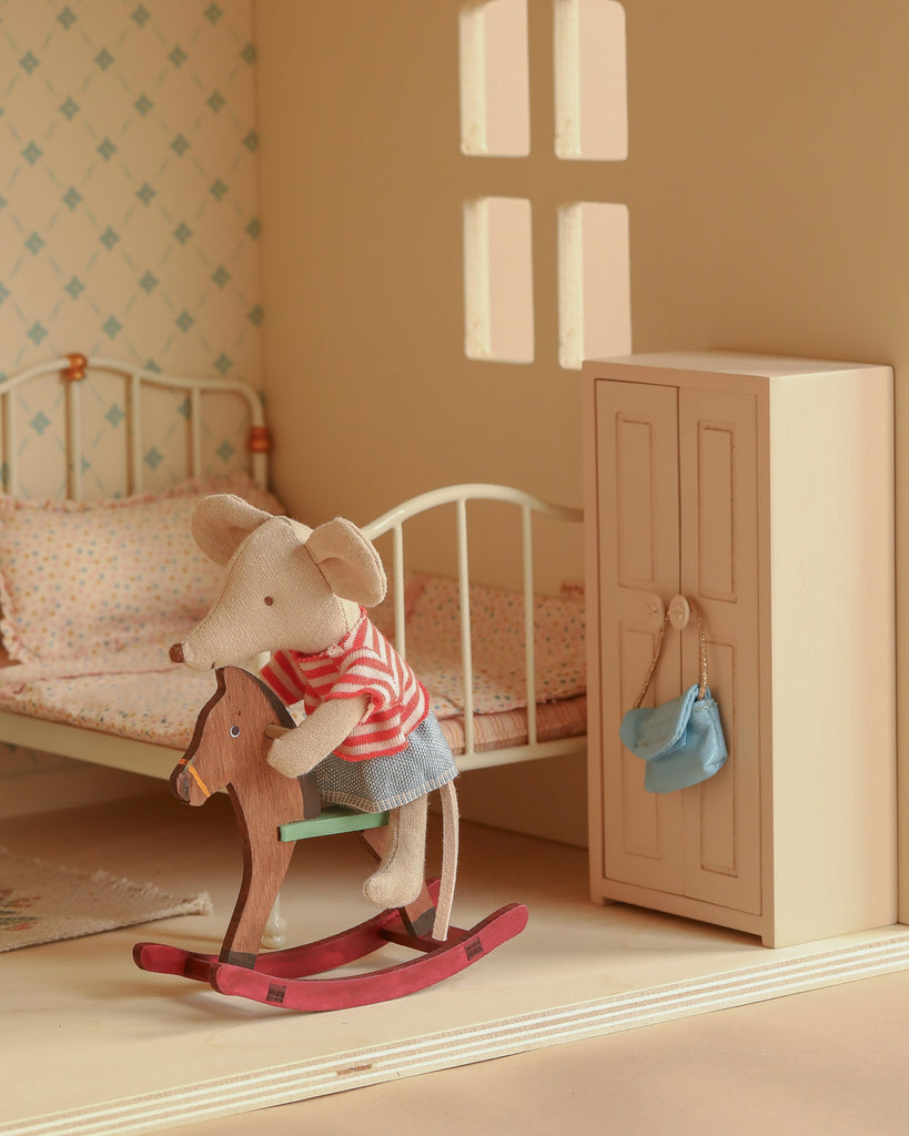 A Maileg Fully Furnished Dollhouse Set + Gift With Purchase features a stuffed mouse in a red striped shirt and blue shorts riding a wooden rocking horse in a cozy bedroom with bed, wardrobe, rug, and natural window light.