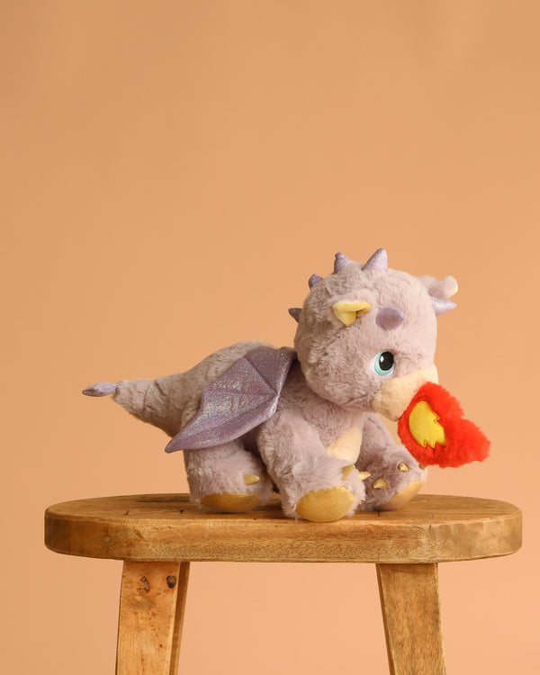 The Olli Ella Dinkum Dragon - Flare - Lavender, a purple plush dragon with sparkly wings and yellow horns, sits on a wooden stool holding a red and orange flame in its magnetic mouth against a plain peach background.
