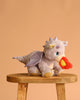 The Olli Ella Dinkum Dragon - Flare - Lavender, a purple plush dragon with sparkly wings and yellow horns, sits on a wooden stool holding a red and orange flame in its magnetic mouth against a plain peach background.