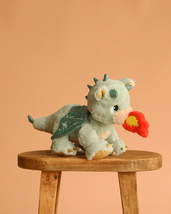 The Olli Ella Dinkum Dragon - Blaze - Green, a plush dragon with shiny wings and spikes, sits on a wooden stool. Its magnetic mouth holds a felt red-orange flame, set against a plain peach background.