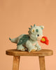 The Olli Ella Dinkum Dragon - Blaze - Green, a plush dragon with shiny wings and spikes, sits on a wooden stool. Its magnetic mouth holds a felt red-orange flame, set against a plain peach background.