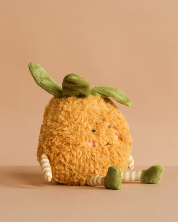 The MON AMI Pina Pineapple Plush Toy features a fuzzy orange body, green leafy top, striped arms and legs, and blushing cheeks—an adorable plush sitting on a beige surface, perfect as a gift for all ages.
