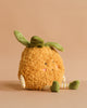 The MON AMI Pina Pineapple Plush Toy features a fuzzy orange body, green leafy top, striped arms and legs, and blushing cheeks—an adorable plush sitting on a beige surface, perfect as a gift for all ages.