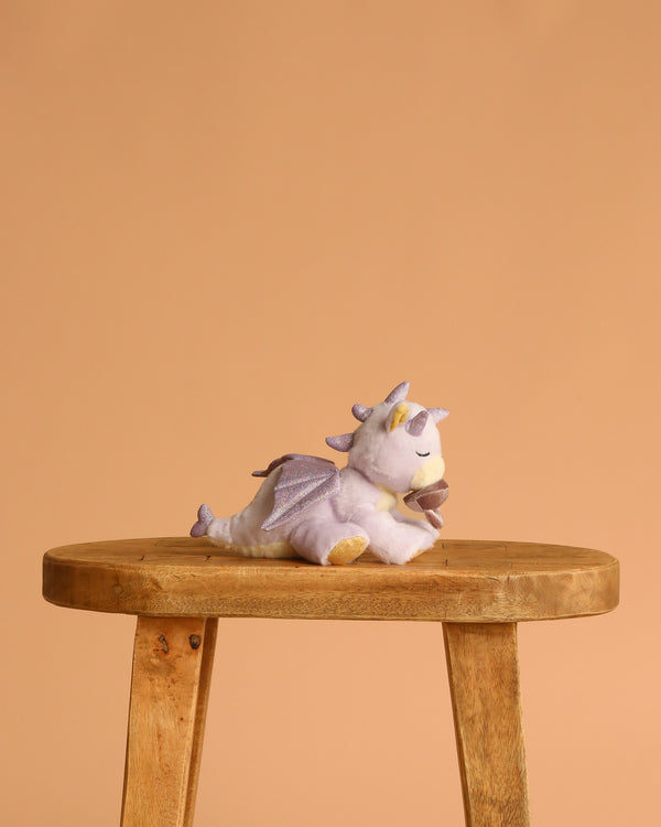 The Olli Ella Binky Dragon - Misty - Lavender, a small plush dragon with purple wings and a removable magnetic pacifier, sits on a simple wooden stool against a plain beige background.