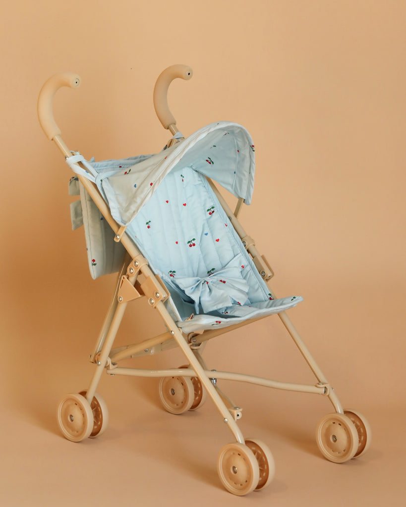 The Konges Sloejd Zoe Bow Doll Stroller - Cherry Blue features a light blue canopy and seat cover adorned with small red and black designs. It includes beige handles and frame, double wheels, and a foldable shade for convenience. The stroller's minimalist design is accentuated by its plain, soft beige background. No baby inside.