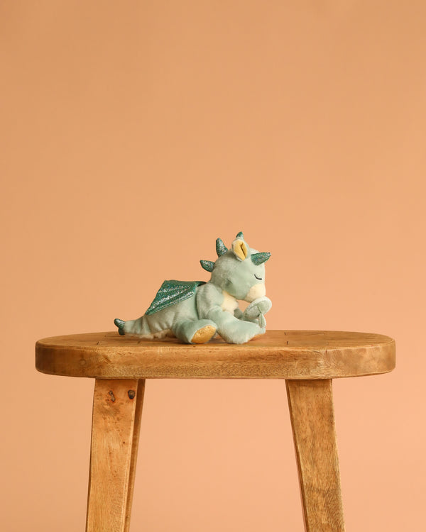 The Olli Ella Binky Dragon - Volt - Green, featuring a yellow horn and wings, rests on its side atop a wooden stool against a plain peach background for an adorable cuddly toy display.