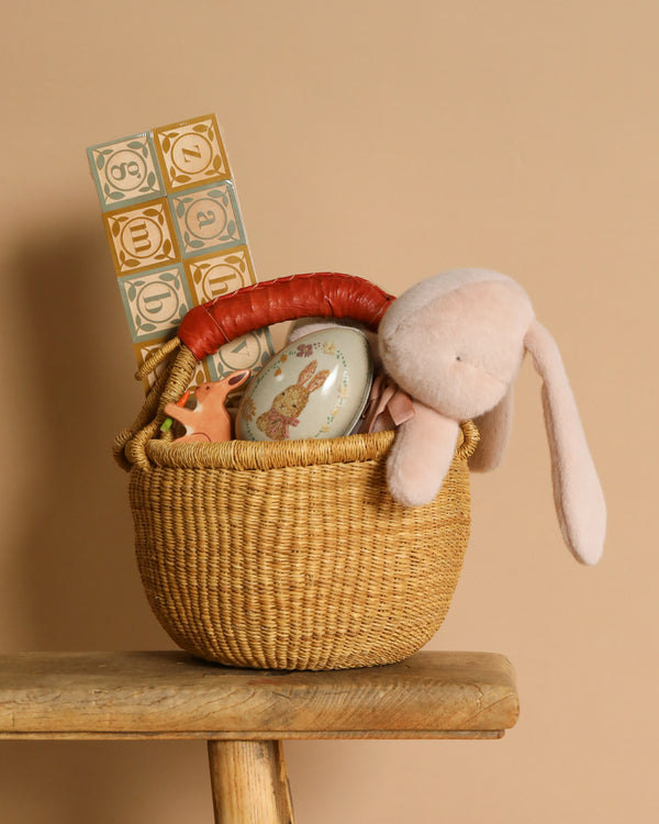 The Easter Basket Set from Bolgatanga, Ghana, features a handmade basket on a wooden stool. It holds stuffed toys, an oval tin, and a block puzzle. A plush long-eared bunny and a charming wooden toy complement the soft beige background of intricately woven elephant grass.