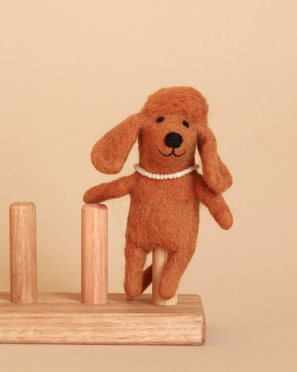 A handmade, brown felt Poodle Dog Finger Puppet with droopy ears and a white beaded necklace stands upright on one leg on a simple wooden toy stand with three vertical pegs. The background is a plain beige, complementing the warm tones of the felt and wood.
