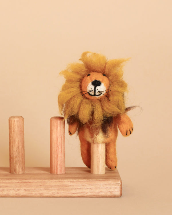A small, Fluffy Lion Finger Puppet with a fluffy mane stands on a wooden peg board against a beige background. Handmade in Nepal, the puppet has a cheerful expression and is positioned atop one of the vertical wooden pegs. Perfect as a storytelling prop, it brings joy and imagination to any tale.