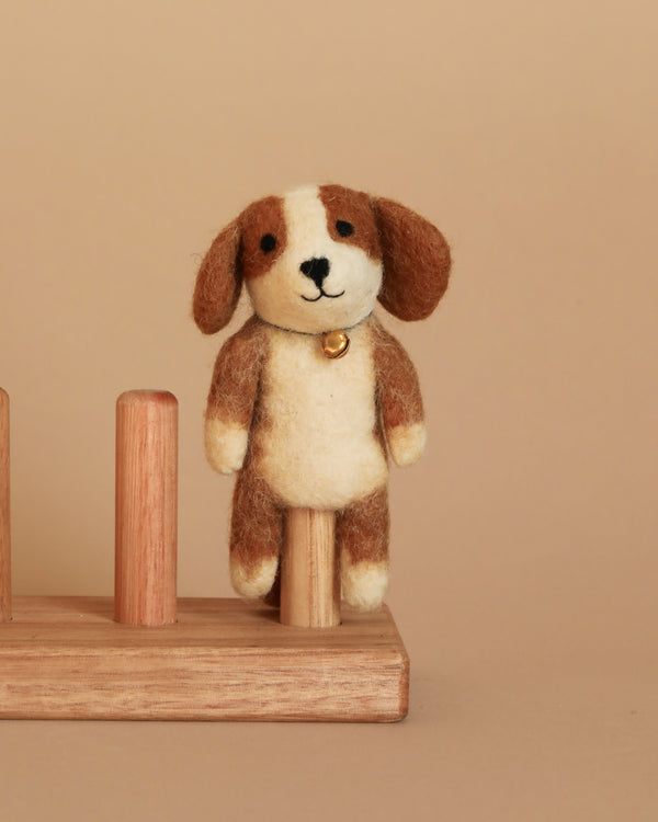 The Cavalier King Charles Spaniel Dog Finger Puppet is a small, handcrafted felt dog with brown ears and spots, standing on a wooden peg. It features needle felt details, a stitched smile, black eyes, and wears a tiny golden bell around its neck. The background is a solid beige color.
