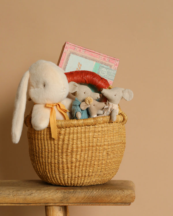 The Maileg Easter Basket Set, featuring a stuffed white bunny with an orange ribbon, two gray plush mice, and a red and pink book, nestles in a woven handmade basket on a wooden stool against a beige background.