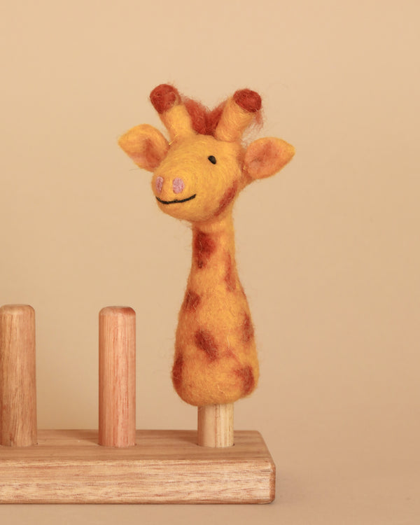 The Giraffe Finger Puppet boasts a long neck and brown spots, highlighted by a pink nose and a smiling face. It is mounted on a wooden stand that has three empty pegs, against a plain beige background.