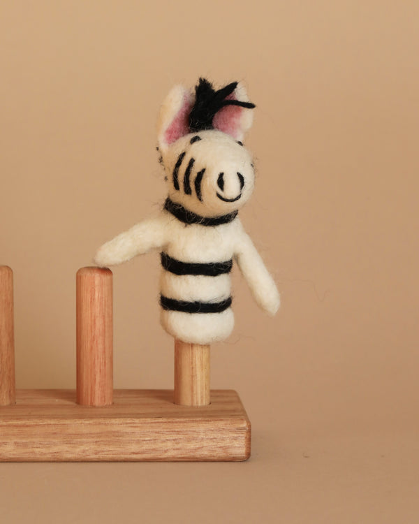The Zebra Finger Puppet, a handmade piece featuring black and white stripes, a black yarn mane, and pink inner ears, is elegantly displayed on a wooden stand with three pegs against a beige background. The middle peg cradles the puppet—ideal for enthusiasts of wild animals.