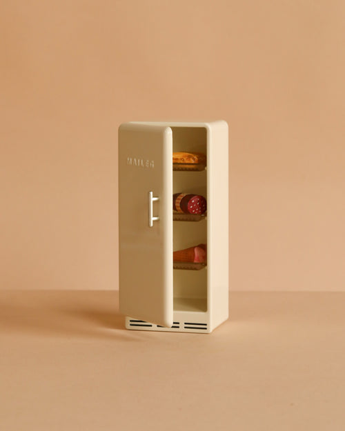 The Maileg Metal Fridge, Mouse - Off White features an open door showing tiny food items on its shelves, set against a plain beige background—ideal for any dollhouse or Maileg Mouse hole Farmhouse.