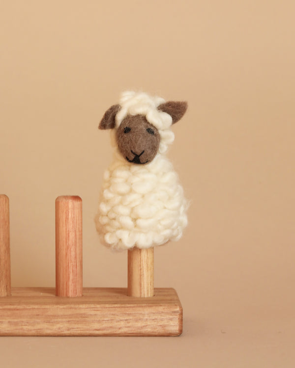 A charming Fluffy Sheep Finger Puppet, crafted by hand in Nepal from 100% wool, features a dark face and ears and sits gracefully on a wooden peg. The backdrop is a gentle beige color that enhances the natural hues of both the puppet and the peg.