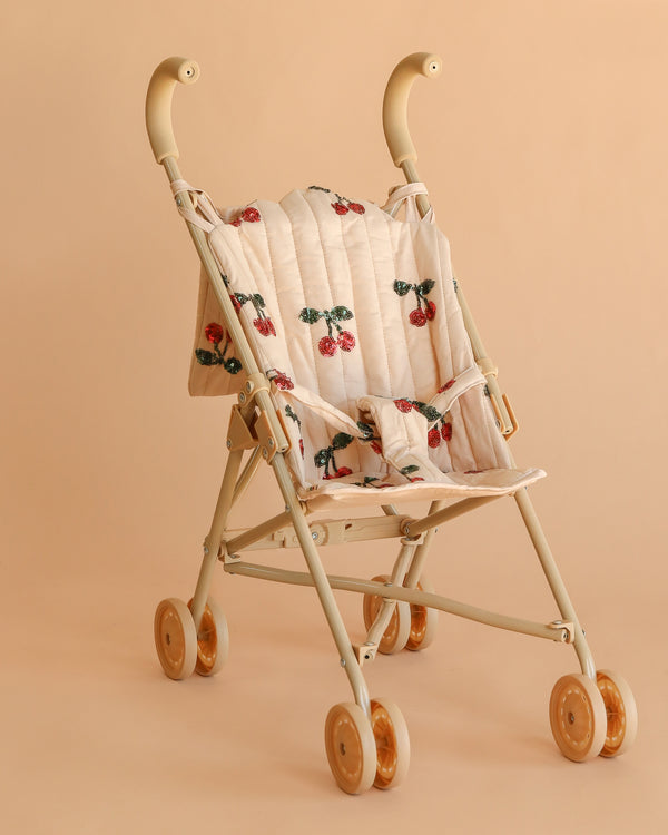 The Konges Sloejd Tulle Doll Stroller - Cherry Sequin features a beige foldable frame, quilted cherry-patterned seat, rounded handles, and four sets of double wheels, shown here against a matching beige background.