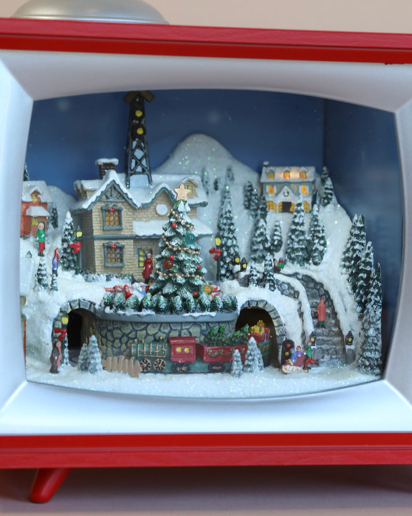 A miniature snowy Christmas village displayed inside a red and white window frame, featuring decorative houses, a Christmas Music TV With Depot And Train, and trees, all covered in snow.